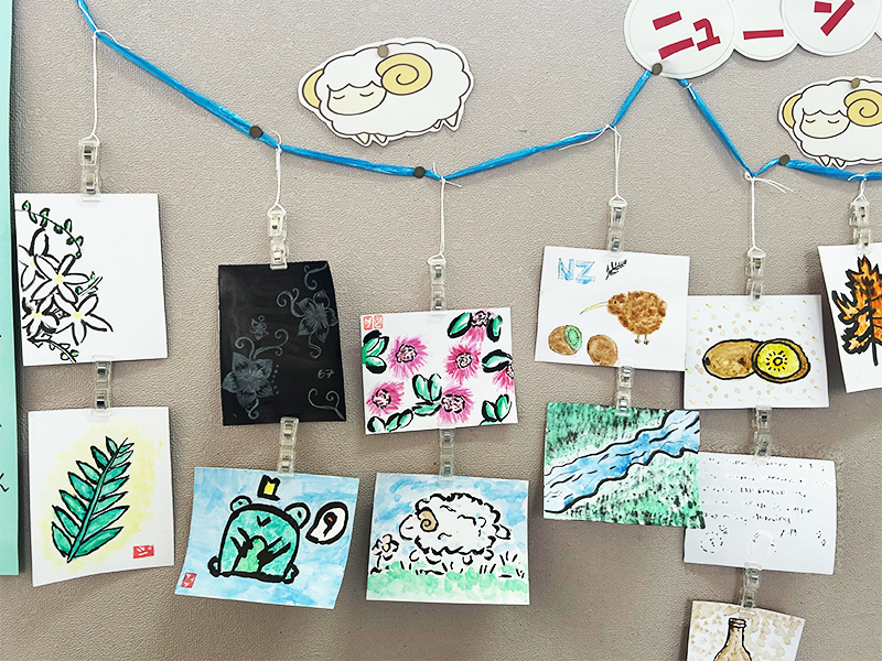左の写真をフォーカスしたニュージーランドらしい絵柄の作品が並んでいる。絵手紙には、花や川などの自然、羊やキーウィなどの動物、キウイフルーツ等の果物が描かれている。