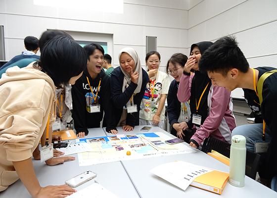 楽しげに笑いながら、地域オリエンテーリングについてテーブルの周りを囲んで立って話し合ういろいろな国・地域の高校生