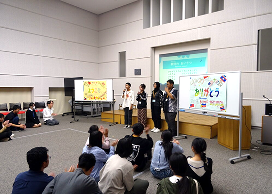 天井が高く広い室内で、中央奥のスクリーン前に4人のいろいろな国・地域の生徒が立って、日本の高校生に歓迎の気持ちを伝えている。左のホワイトボードには「ようこそ」、右のホワイトボードには「ありがとう」とあり、その前にはたくさんの生徒が座って拍手している。