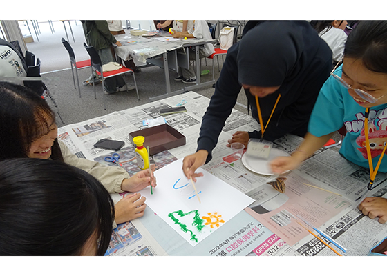 新聞紙を敷いたテーブルの上でインドネシア、ベトナム、日本の高校生が相談しながら、色のついた砂を画用紙にふりかけて絵を描いている。