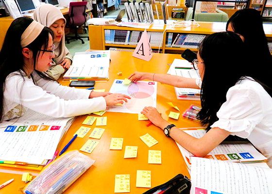 テーブルを４人の大学生が囲みながら、付箋に書いたことを読み上げながら、ベン図が印刷された用紙の上に貼る作業をしている。