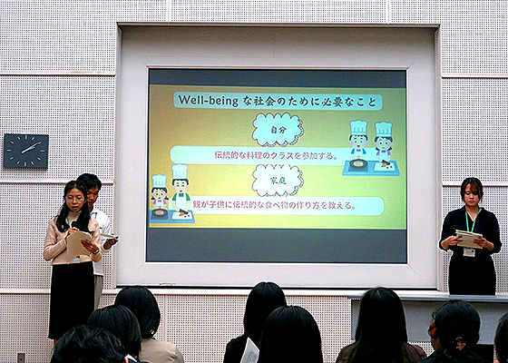 大きなスクリーンの左に二人、右に一人の学生が立ち、「Well-beingな社会のために必要なこと」について発表している。