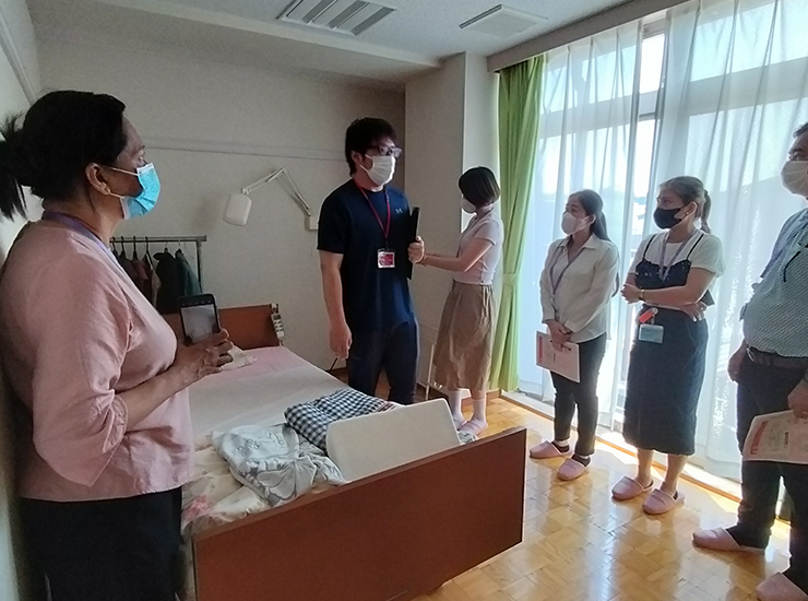 白いレースのカーテンがかかる大きな窓を背に、利用者の実際の居室で介護スタッフの説明を並んで聞いている研修参加者の写真