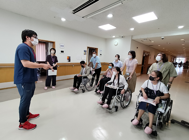 広い空間で、車いす体験をしながら介護スタッフの説明を聞く研修参加者たちの写真