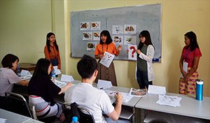 教壇でイラストカードや「？」が書かれたカードを見せながらミャンマー語を教える先生役の受講生と、それを聞く学生の写真