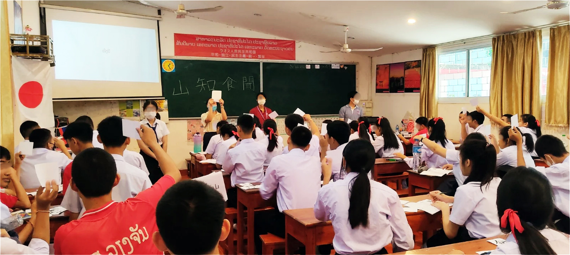 教室で白いシャツを着た生徒たちが漢字カードを手に掲げている。