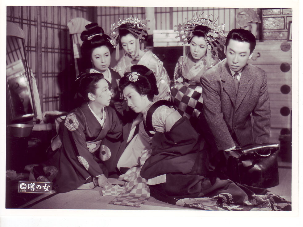 映画『噂の女』の1シーン。1950年代の屋内に豪華な髪飾りや衣装を身にまとった女性たち5人と、スーツを着た男性が1人写っている。