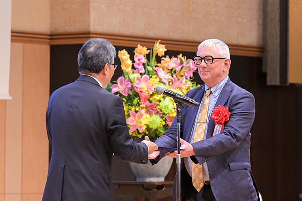 舞台上で賞を手渡すJFの理事長と、受け取る受賞者のアップ 後ろに大きな演台花が見えている