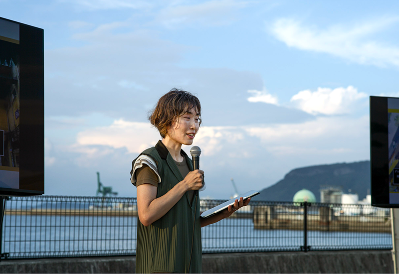 マイクを手に話をする女性、背景に海が見えている