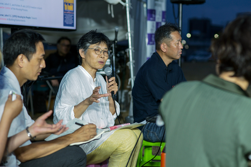 マイクを持った女性がプラスチックの椅子に座って話している。