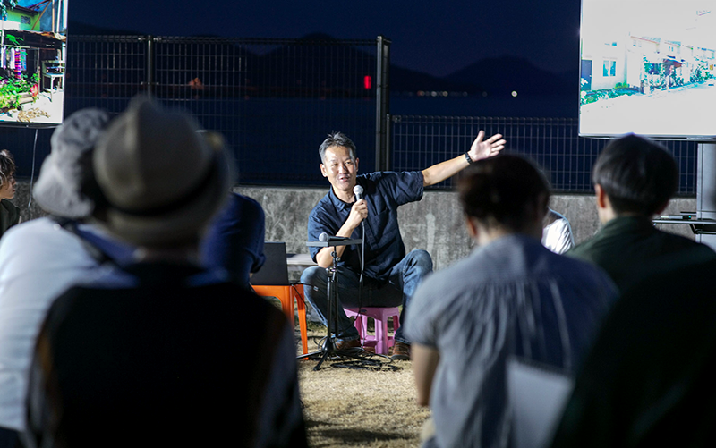 夜の海を前に、椅子に座り片手を広げて集まった人々に向かってマイクをもって話す男性。