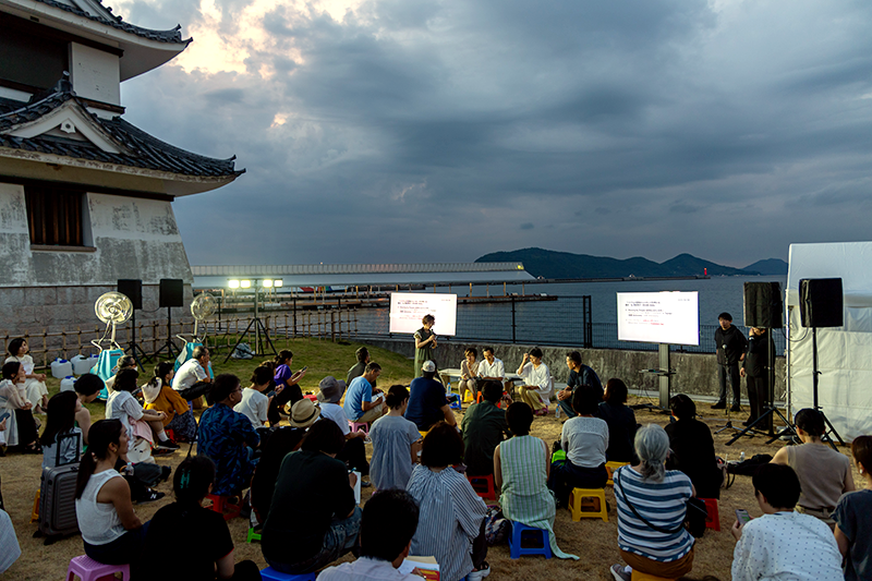 背景に海の見える会場の様子。人々が集まり、前に数人の登壇者やスタッフが発言している